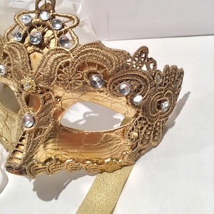 May include: A gold masquerade mask with intricate lace detailing and rhinestones. The mask has a gold ribbon attached.