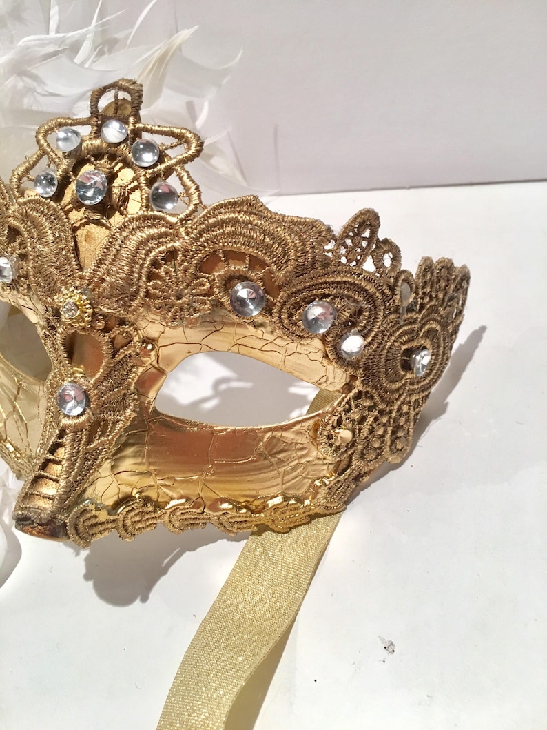May include: A gold masquerade mask with intricate lace detailing and rhinestones. The mask has a gold ribbon attached.