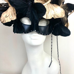 Puede incluir: Una m&aacute;scara negra de carnaval con detalles de mariposas doradas y negras. La m&aacute;scara est&aacute; adornada con plumas y pedrer&iacute;a, y se exhibe sobre una cabeza de maniqu&iacute; blanca. Dos hebras de cuentas cuelgan de la m&aacute;scara.