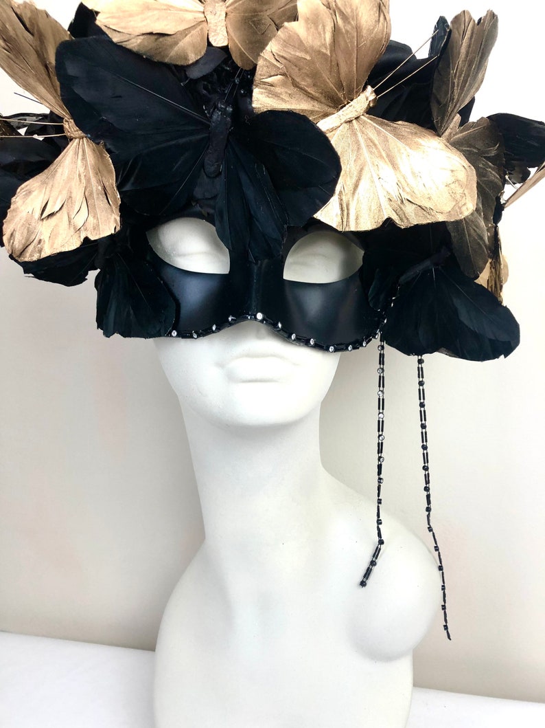 Puede incluir: Una m&aacute;scara negra de carnaval con detalles de mariposas doradas y negras. La m&aacute;scara est&aacute; adornada con plumas y pedrer&iacute;a, y se exhibe sobre una cabeza de maniqu&iacute; blanca. Dos hebras de cuentas cuelgan de la m&aacute;scara.