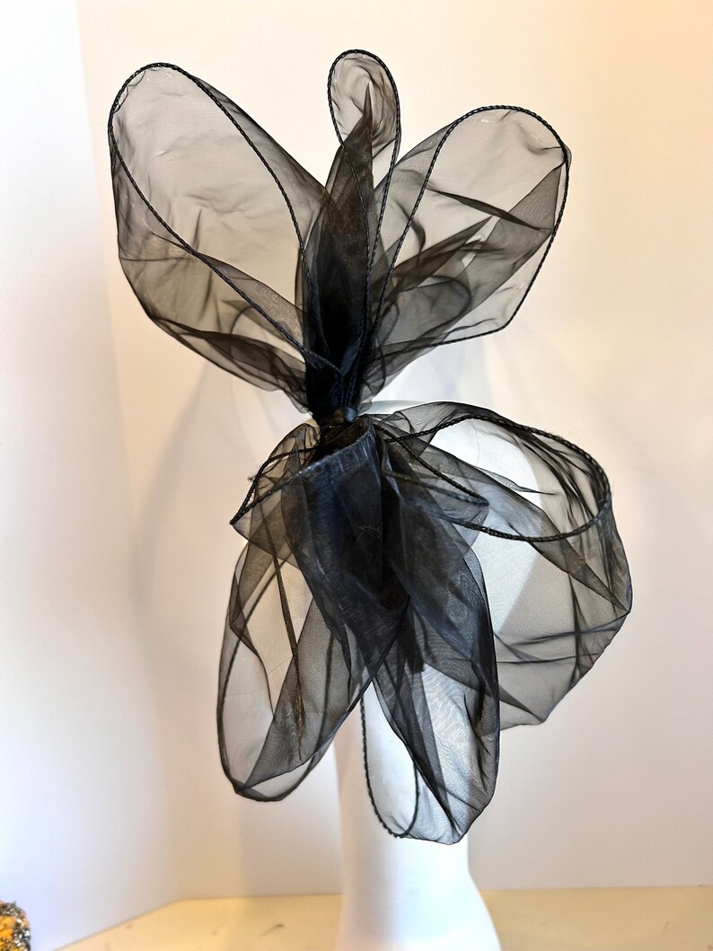 May include: A large black sheer bow with a wired edge. The bow is tied in a knot and is ready to be attached to a hat or other accessory.