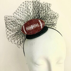 May include: A decorative headpiece featuring a brown football design with white accents. The football is embellished with rhinestones and set against a black net and a black headband. The headpiece is designed for special events.