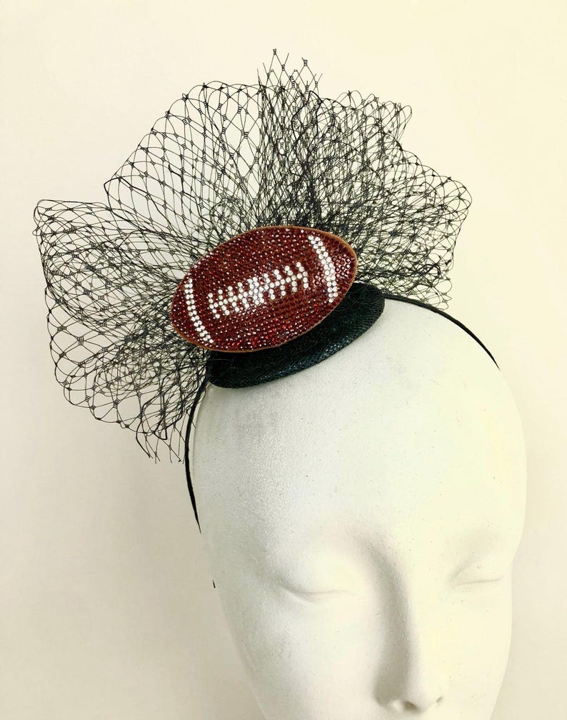 May include: A decorative headpiece featuring a brown football design with white accents. The football is embellished with rhinestones and set against a black net and a black headband. The headpiece is designed for special events.