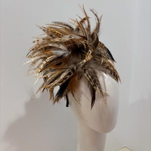 K&ouml;nnte beinhalten: Ein braunes und schwarzes Federhaarband mit einem gro&szlig;en, flauschigen Federakzent. Das Haarband wird auf einem wei&szlig;en Mannequin-Kopf getragen.