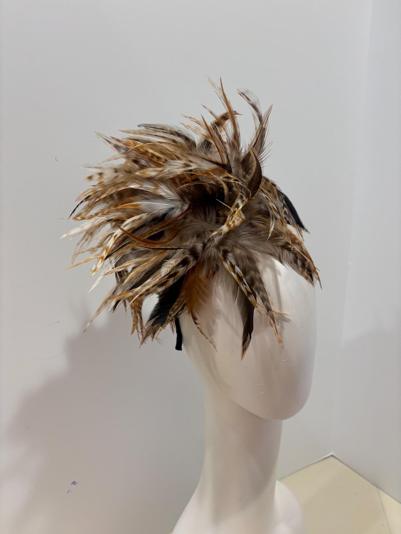 K&ouml;nnte beinhalten: Ein braunes und schwarzes Federhaarband mit einem gro&szlig;en, flauschigen Federakzent. Das Haarband wird auf einem wei&szlig;en Mannequin-Kopf getragen.