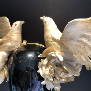 May include: A black headpiece with two gold-colored dove figurines with outstretched wings. The headpiece is adorned with gold-colored flowers and a gold-trimmed band. The background is black.