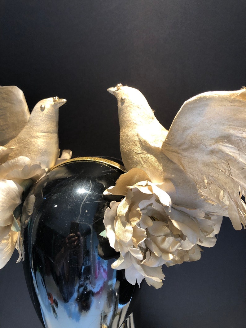 May include: A black headpiece with two gold-colored dove figurines with outstretched wings. The headpiece is adorned with gold-colored flowers and a gold-trimmed band. The background is black.