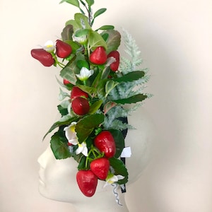 以下が含まれることがあります： 鮮やかな赤いイチゴ、白い花、緑の葉をあしらったヘッドピース。装飾的なアレンジメントには、黒いヘッドバンドに取り付けられた霜で覆われた緑の小枝が含まれています。イチゴは深い赤色です。
