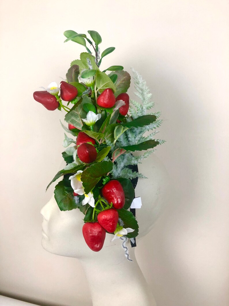 以下が含まれることがあります： 鮮やかな赤いイチゴ、白い花、緑の葉をあしらったヘッドピース。装飾的なアレンジメントには、黒いヘッドバンドに取り付けられた霜で覆われた緑の小枝が含まれています。イチゴは深い赤色です。