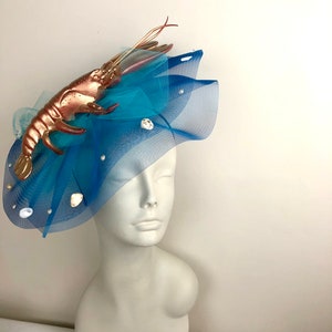 Op de afbeelding: Een blauwe tule fascinator met een gouden kreeft en witte schelpen. De fascinator is op een wit mannequins hoofd.