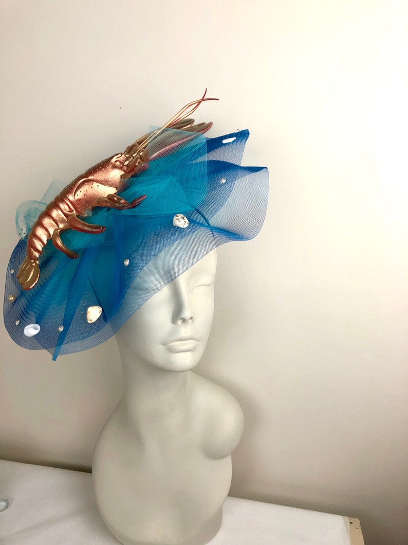 Op de afbeelding: Een blauwe tule fascinator met een gouden kreeft en witte schelpen. De fascinator is op een wit mannequins hoofd.