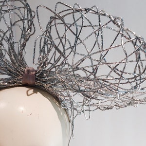 May include: A silver wire headband with a large, intricate bow. The headband is made of thin, interwoven wires that create a delicate and airy look.