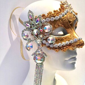 May include: A gold masquerade mask with a silver lace trim and a crystal embellishment. The mask has a long crystal tassel hanging from the bottom.