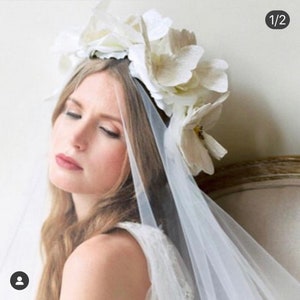 May include: A white bridal veil with a floral crown made of white flowers. The veil is sheer and delicate, and the flowers are arranged in a circle around the bride's head.