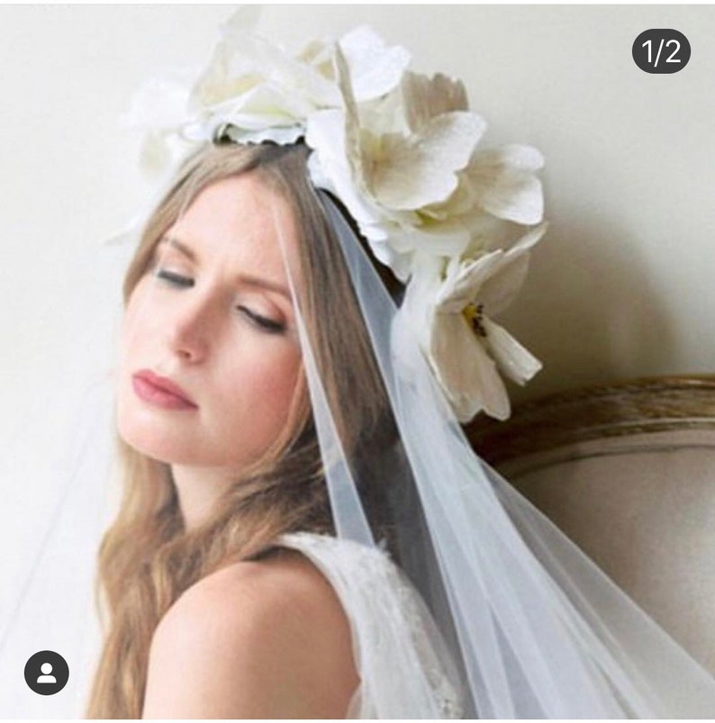 May include: A white bridal veil with a floral crown made of white flowers. The veil is sheer and delicate, and the flowers are arranged in a circle around the bride's head.