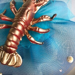 Op de afbeelding: Een gouden kreeftfiguur op een blauwe tule stof met witte schelpen.