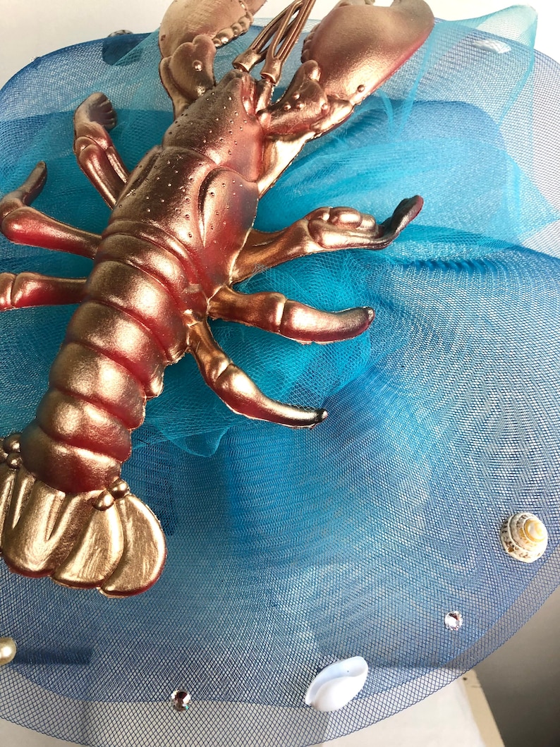 Op de afbeelding: Een gouden kreeftfiguur op een blauwe tule stof met witte schelpen.