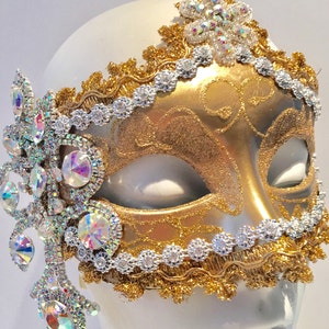 May include: A gold masquerade mask with intricate details and a silver trim. The mask is decorated with iridescent rhinestones and a dangling crystal embellishment.