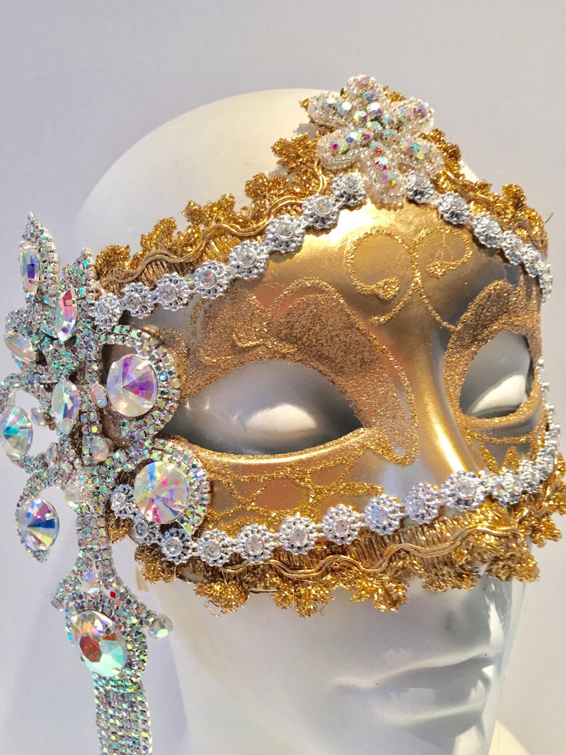 May include: A gold masquerade mask with intricate details and a silver trim. The mask is decorated with iridescent rhinestones and a dangling crystal embellishment.
