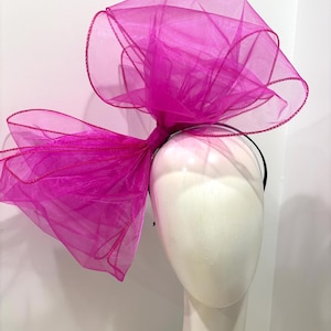 May include: A large, bright pink tulle bow headband. The bow is made of soft, sheer fabric and is tied in a large, dramatic knot. The headband is black and is designed to be worn around the head.