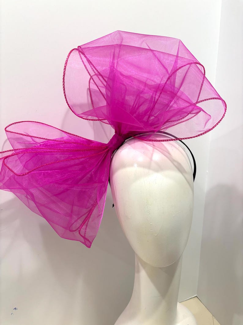 May include: A large, bright pink tulle bow headband. The bow is made of soft, sheer fabric and is tied in a large, dramatic knot. The headband is black and is designed to be worn around the head.