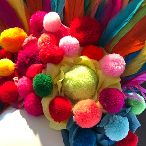 May include: A colorful headpiece with a variety of bright pom-poms and feathers. The pom-poms are in shades of red, pink, yellow, green, and blue. The feathers are in shades of orange, red, pink, purple, and blue.