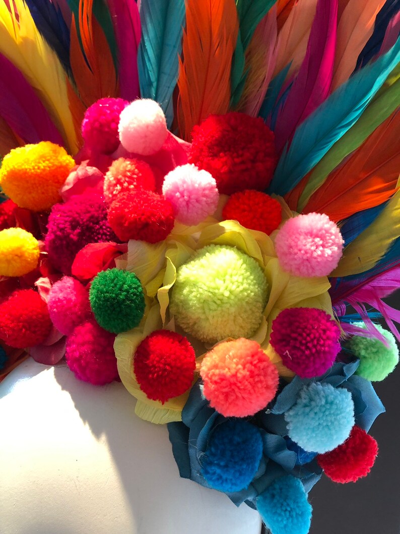 May include: A colorful headpiece with a variety of bright pom-poms and feathers. The pom-poms are in shades of red, pink, yellow, green, and blue. The feathers are in shades of orange, red, pink, purple, and blue.