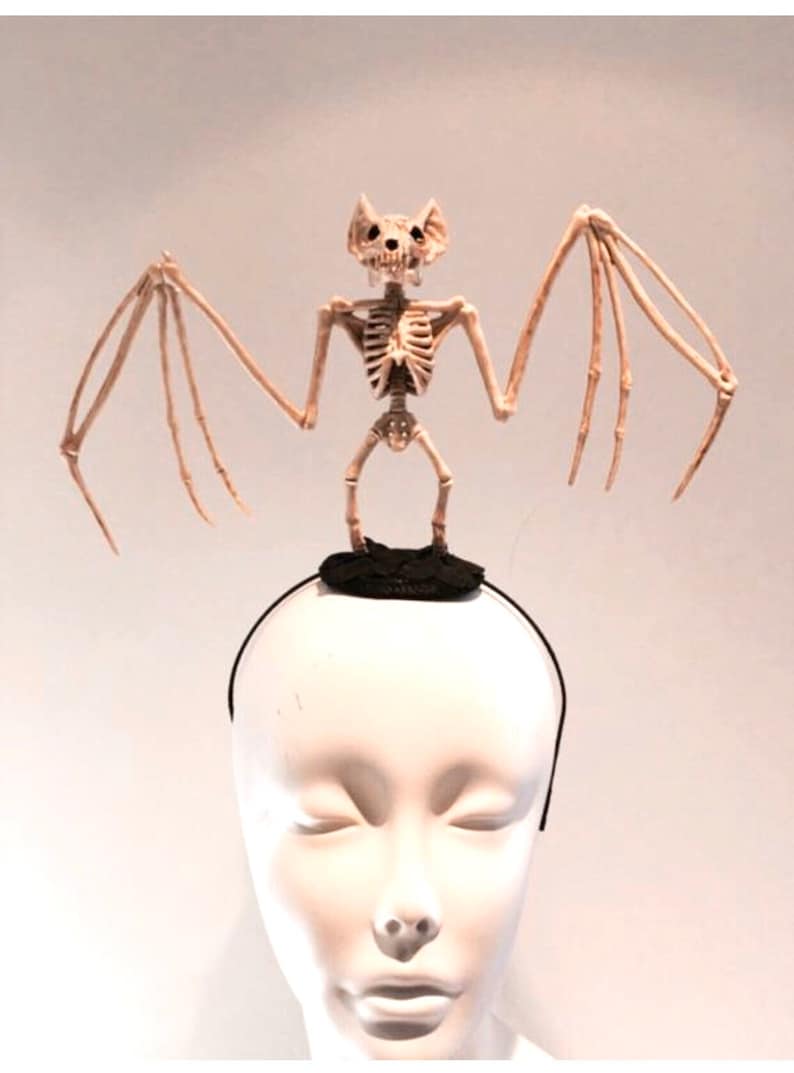 May include: A Halloween-themed headband featuring a skeletal bat figure. The bat is beige and has outstretched wings, perched atop a black, circular base. The headband itself is black, and the display is on a mannequin head.