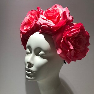 May include: A floral headpiece featuring several large, vibrant pink roses arranged across the top. The artificial flowers are attached to a dark band, creating a crown-like appearance. The headpiece is displayed on a smooth, white mannequin head.