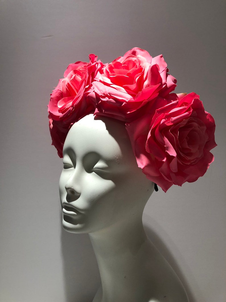 May include: A floral headpiece featuring several large, vibrant pink roses arranged across the top. The artificial flowers are attached to a dark band, creating a crown-like appearance. The headpiece is displayed on a smooth, white mannequin head.
