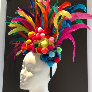 May include: A colorful headband with a large pom-pom flower and feathers. The headband is made of a black elastic band and is decorated with a variety of colors, including red, orange, yellow, green, blue, and purple.