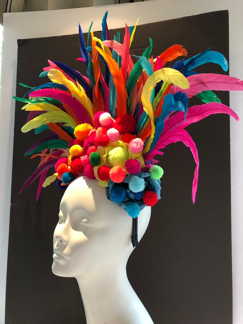May include: A colorful headband with a large pom-pom flower and feathers. The headband is made of a black elastic band and is decorated with a variety of colors, including red, orange, yellow, green, blue, and purple.