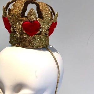 May include: A gold glitter crown headband featuring a series of raised red velvet heart appliqu&eacute;s around the base. The crown has a central spherical finial at the top and is attached to a thin, gold-toned metal headband for secure placement.