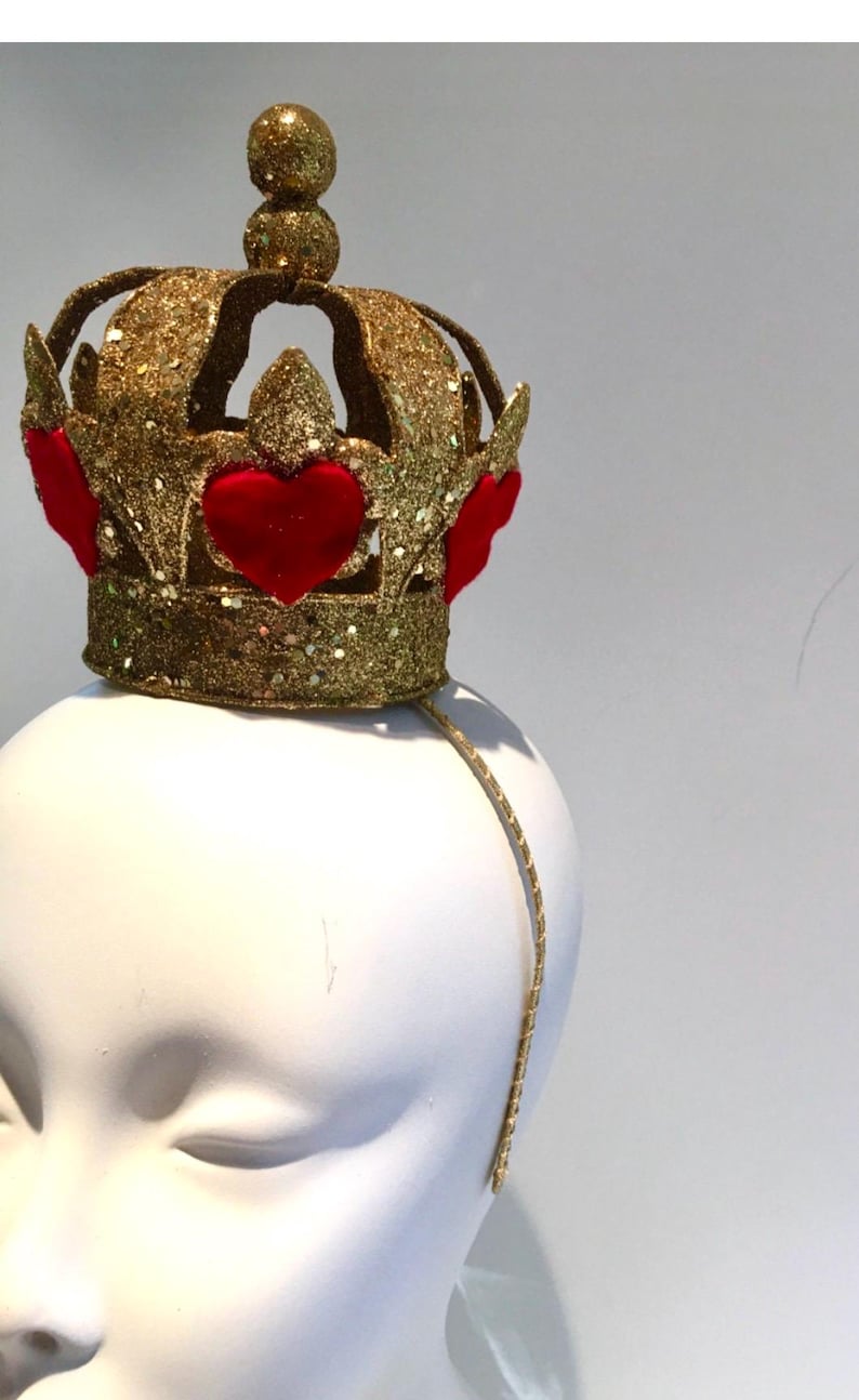 May include: A gold glitter crown headband featuring a series of raised red velvet heart appliqu&eacute;s around the base. The crown has a central spherical finial at the top and is attached to a thin, gold-toned metal headband for secure placement.