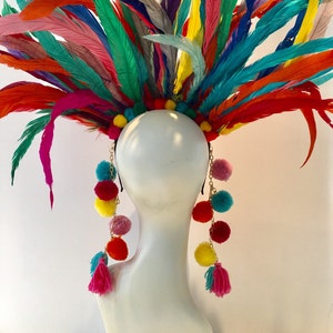 May include: A colorful feather headdress with long strands of pom-poms and tassels hanging down the sides. The headdress is made of a variety of feathers in shades of pink, blue, green, yellow, and orange. The pom-poms are also in a variety of colors, including pink, yellow, red, and blue.