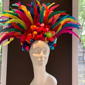May include: A colorful headpiece with a rainbow of feathers and pom-poms. The headpiece is made of bright colors, including red, orange, yellow, green, blue, indigo, and violet. The headpiece is worn on a white mannequin.