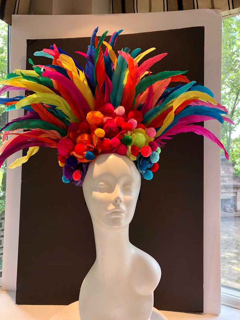 May include: A colorful headpiece with a rainbow of feathers and pom-poms. The headpiece is made of bright colors, including red, orange, yellow, green, blue, indigo, and violet. The headpiece is worn on a white mannequin.