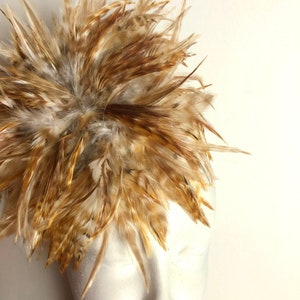 May include: A brown and beige feather headpiece with a fluffy texture. The feathers are arranged in a circular shape and are attached to a white mannequin head.