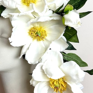 K&ouml;nnte beinhalten: Nahaufnahme von wei&szlig;en Pfingstrosen mit gelben Zentren und gr&uuml;nen Bl&auml;ttern. Die Bl&uuml;tenbl&auml;tter sind weich und geschichtet, mit einer realistischen Textur. Die Blumen sind in einem kaskadierenden Design angeordnet, was auf ein Blumenarrangement oder ein Haaraccessoire hindeutet.