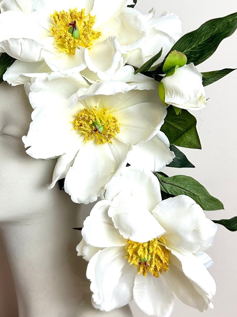 K&ouml;nnte beinhalten: Nahaufnahme von wei&szlig;en Pfingstrosen mit gelben Zentren und gr&uuml;nen Bl&auml;ttern. Die Bl&uuml;tenbl&auml;tter sind weich und geschichtet, mit einer realistischen Textur. Die Blumen sind in einem kaskadierenden Design angeordnet, was auf ein Blumenarrangement oder ein Haaraccessoire hindeutet.