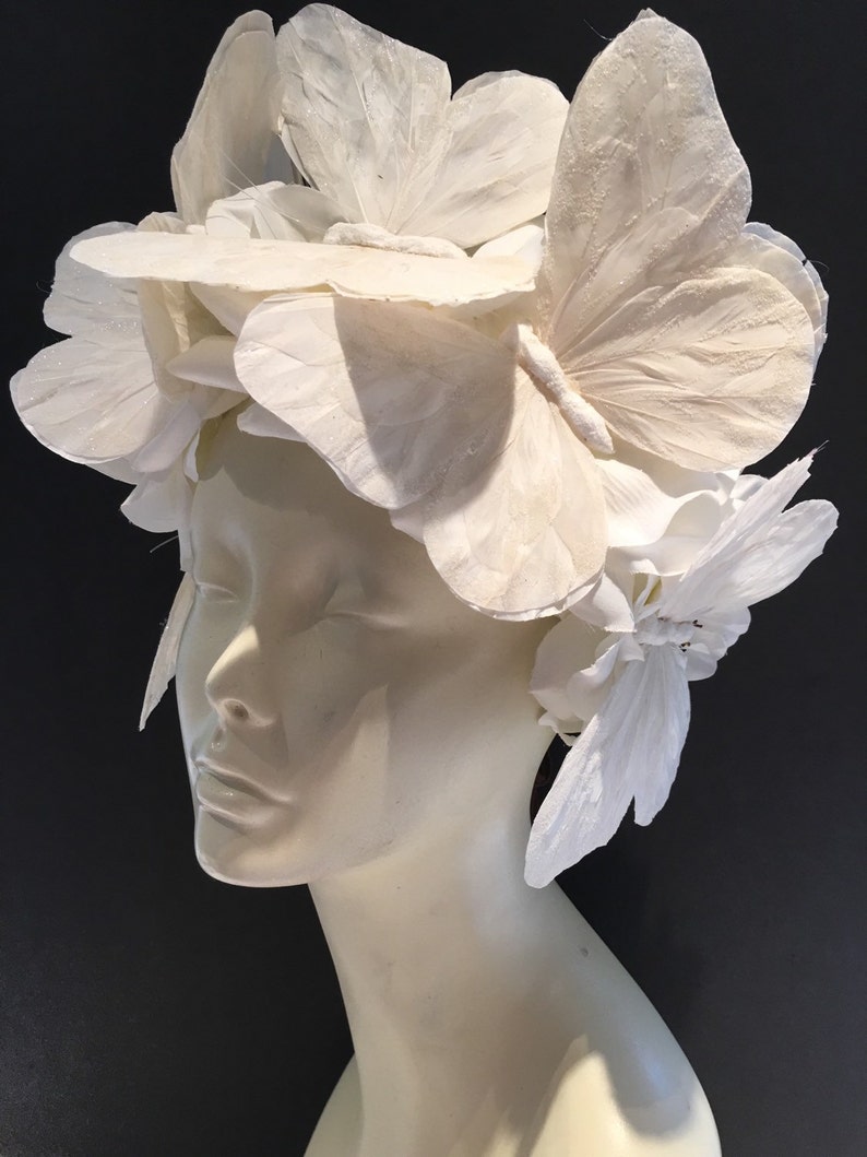 May include: A white headpiece made of paper butterflies and flowers. The headpiece is worn on a white mannequin head.