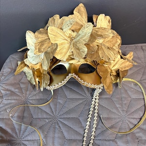 Puede incluir: Una m&aacute;scara dorada para un baile de m&aacute;scaras con dise&ntilde;o de mariposa. La m&aacute;scara presenta una corona de mariposas y un ribete de joyas. Cintas doradas y una cadena de joyas cuelgan de la m&aacute;scara.