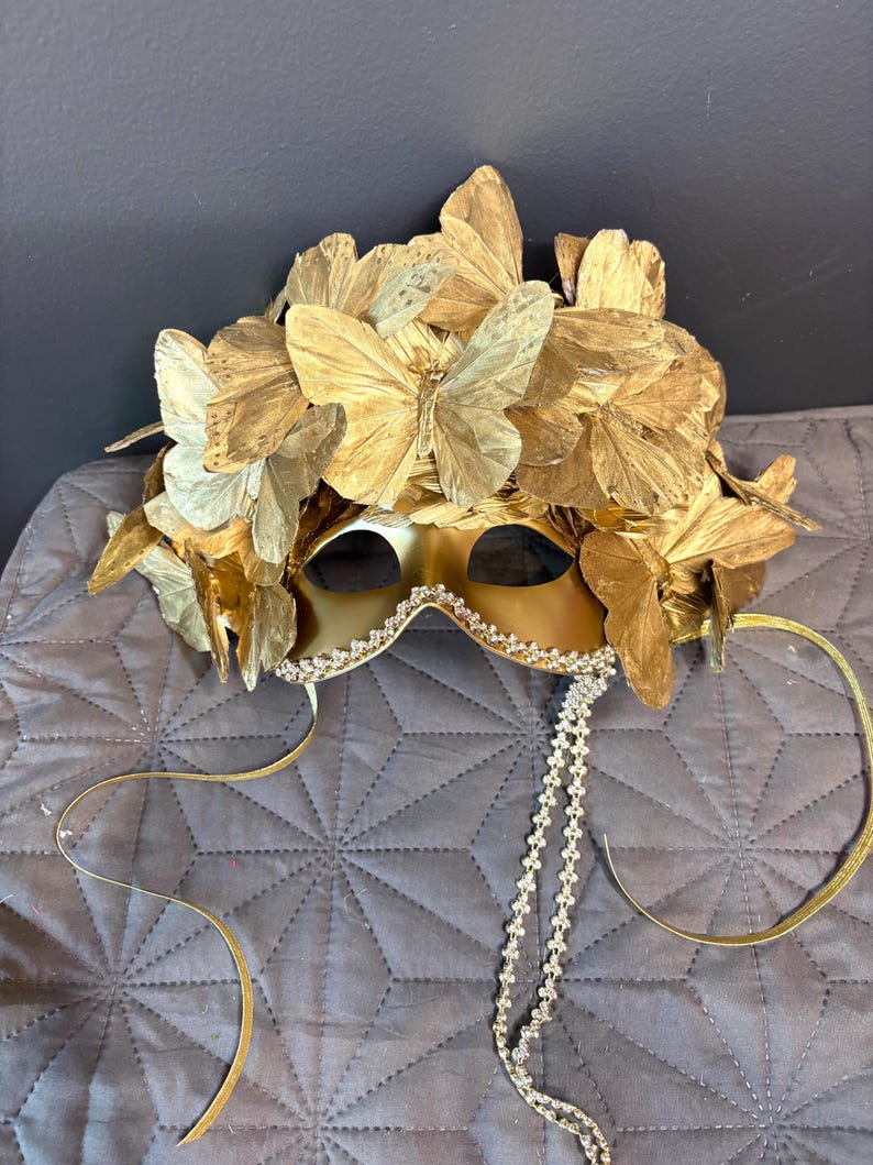 Puede incluir: Una m&aacute;scara dorada para un baile de m&aacute;scaras con dise&ntilde;o de mariposa. La m&aacute;scara presenta una corona de mariposas y un ribete de joyas. Cintas doradas y una cadena de joyas cuelgan de la m&aacute;scara.