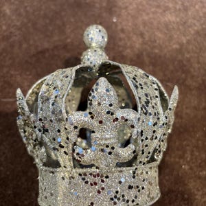 May include: A silver crown ornament covered in fine glitter, featuring a fleur-de-lis design on the front and a small spherical topper. The structure is composed of curved metal bands that form an open, circular shape with a flat base.