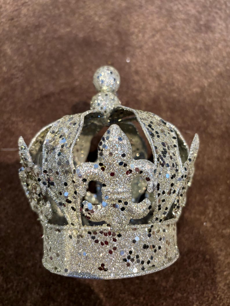 May include: A silver crown ornament covered in fine glitter, featuring a fleur-de-lis design on the front and a small spherical topper. The structure is composed of curved metal bands that form an open, circular shape with a flat base.