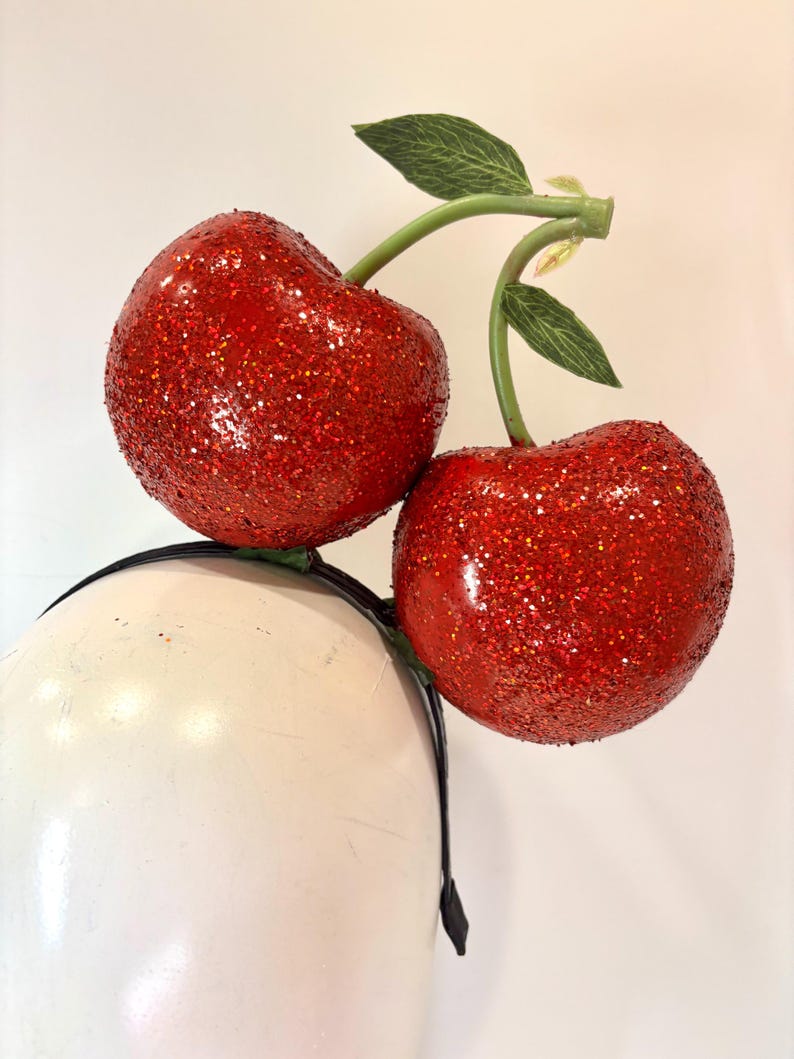 K&ouml;nnte beinhalten: Ein festliches Stirnband mit zwei gro&szlig;en, glitzernden roten Kirschen. Die Kirschen haben gr&uuml;ne Stiele und Bl&auml;tter und sind an einem schwarzen Stirnband befestigt. Die Kirschen sind leuchtend rot und mit Glitzer bedeckt.