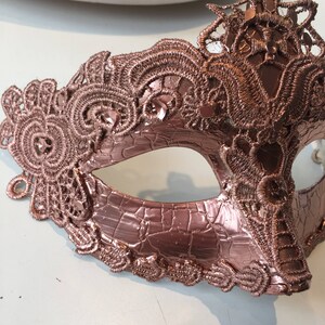 May include: A rose gold masquerade mask with intricate lace detailing. The mask is made of a textured material and has a delicate, feminine design.