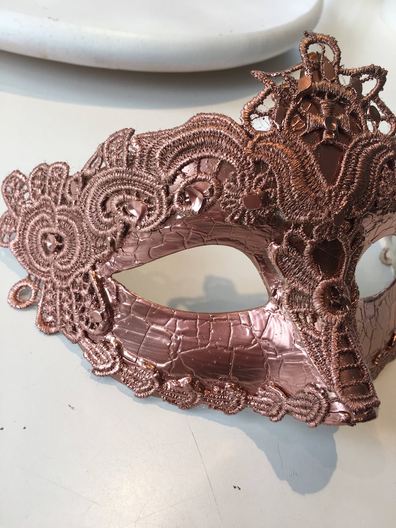 May include: A rose gold masquerade mask with intricate lace detailing. The mask is made of a textured material and has a delicate, feminine design.