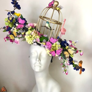 Puede incluir: Un tocado caprichoso con una jaula dorada, un flamenco rosa y mariposas de colores. El tocado está adornado con una variedad de flores y pequeños pájaros, creando un diseño vibrante y artístico. El tocado se exhibe en una cabeza de maniquí blanca.