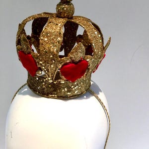 May include: A gold glitter crown headband featuring a series of raised red heart-shaped accents around the base. The crown has a central spherical finial at the top and is attached to a thin, matching gold glitter-covered headband frame.